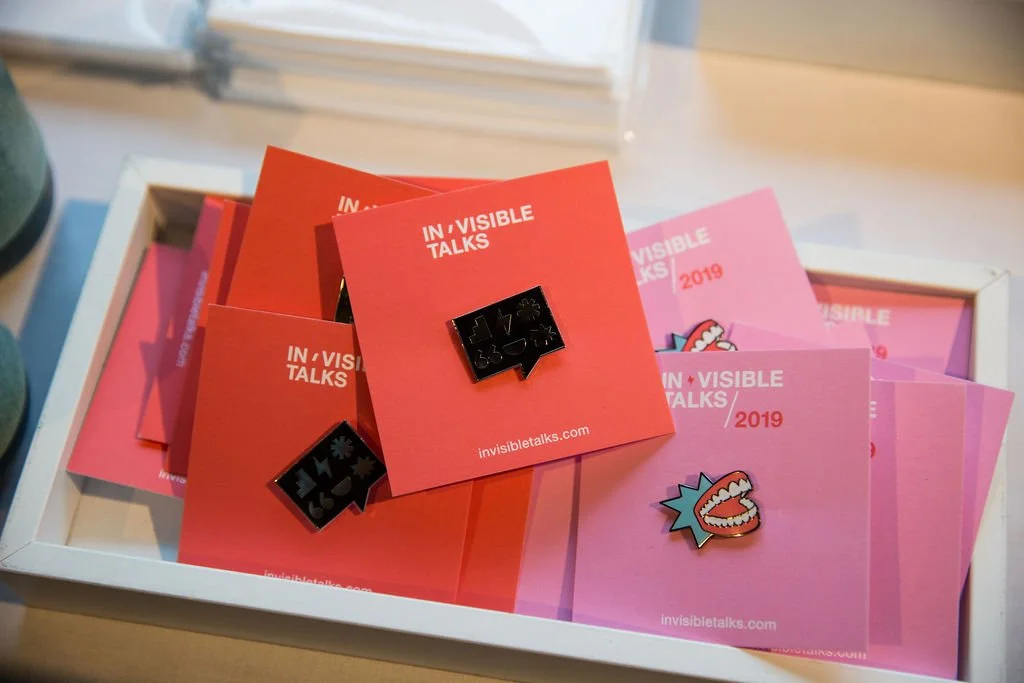 A white tray contains several pink and red cards with white text reading "INVISIBLE TALKS 2019" and a website link. Some cards feature colorful enamel pins, including one with a mouth and teeth, and others with chat bubble icons.