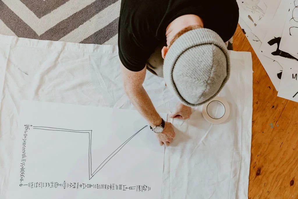 Overhead view of a person wearing a light gray beanie, black shirt, and wristwatch, drawing a graph with black marker on large white paper on the floor. The paper has a line graph with labeled axes and some handwritten notes or labels at the bottom. 