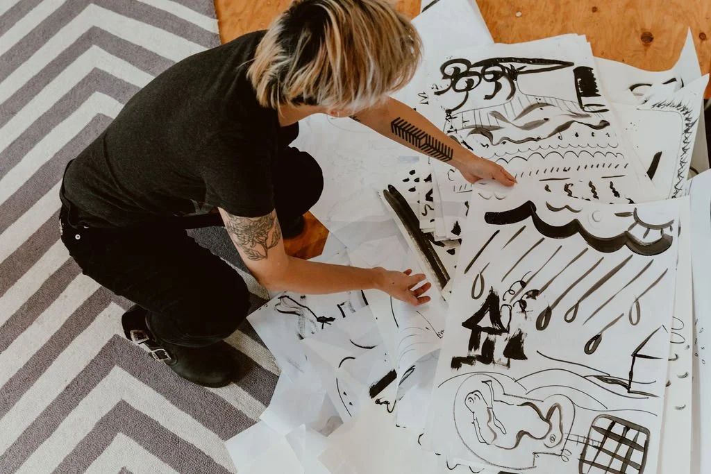 Person with short blonde hair and tattoos on their arm, wearing black clothing, squatting and pointing at large sheets of paper with black marker drawings on the floor.