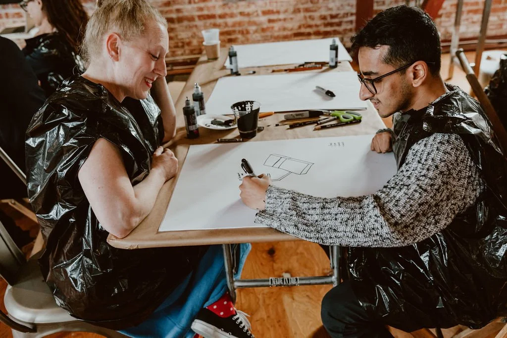 Two people, a woman and a man, sitting at a table with drawing materials, drawing a 3D shape on a large sheet of paper, both smiling, in an art studio with brick walls.