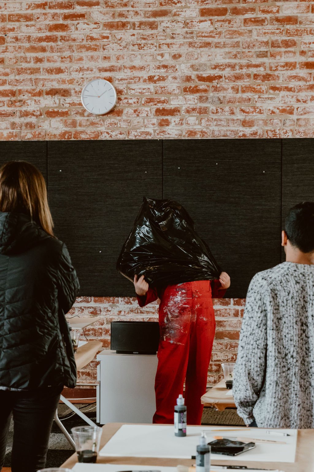 A person wearing red clothes is using a large black sheet to cover their head and face, while two other people are observing.