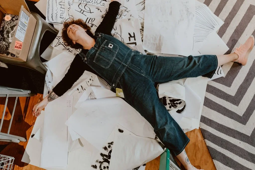 Person lying on their back on a cluttered table covered with papers, some with sketches and writing, surrounded by various objects including a chair and a box, with a striped rug on the floor.