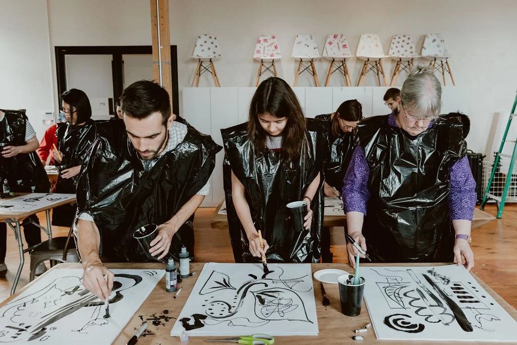 People painting abstract black ink art on paper at tables in an art class.