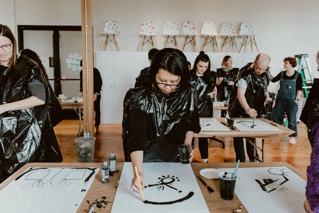 People participating in a group painting activity, with large sheets of paper on tables, paintbrushes, and black paint, in a room decorated with lamps on the wall.