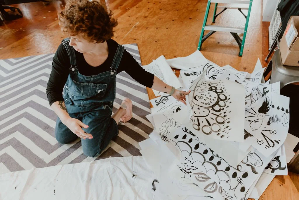 A person with curly hair kneels on a rug and looks at collected black marker drawings on white paper spread across a table and floor.