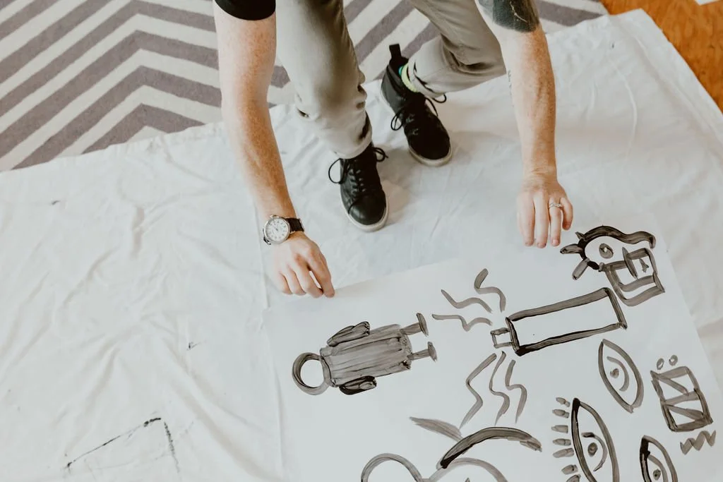 A person is kneeling on a white sheet with a drawing of a person, a fire-hydrant, a streetlamp, a smiley face, and other abstract shapes painted with black marker.