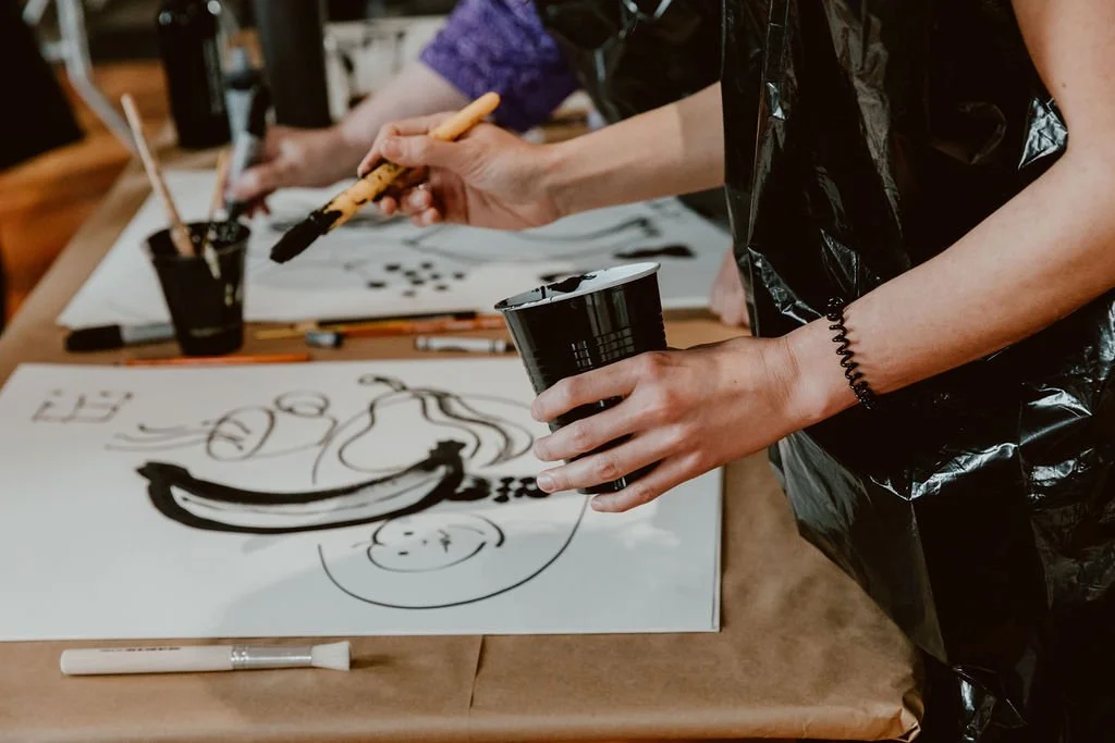 People creating abstract black ink art on paper, using brushes and ink containers, in an art studio.