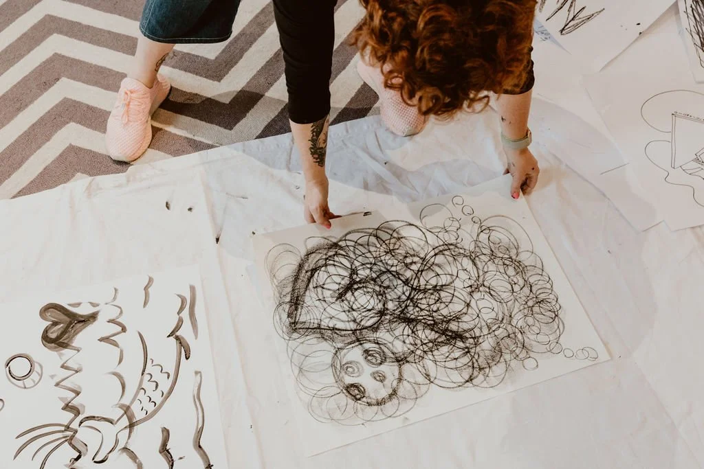 A person with curly red hair creating a scribble drawing of a dog's face on large sheets of paper laid on the floor, surrounded by other sketches and drawings.