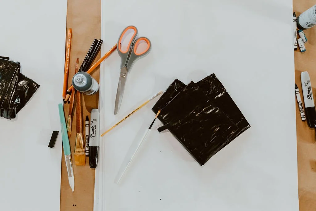 School supplies on a table including scissors, pens, pencils, markers, a glue stick, a white pen, black plastic bags, and a piece of white paper.