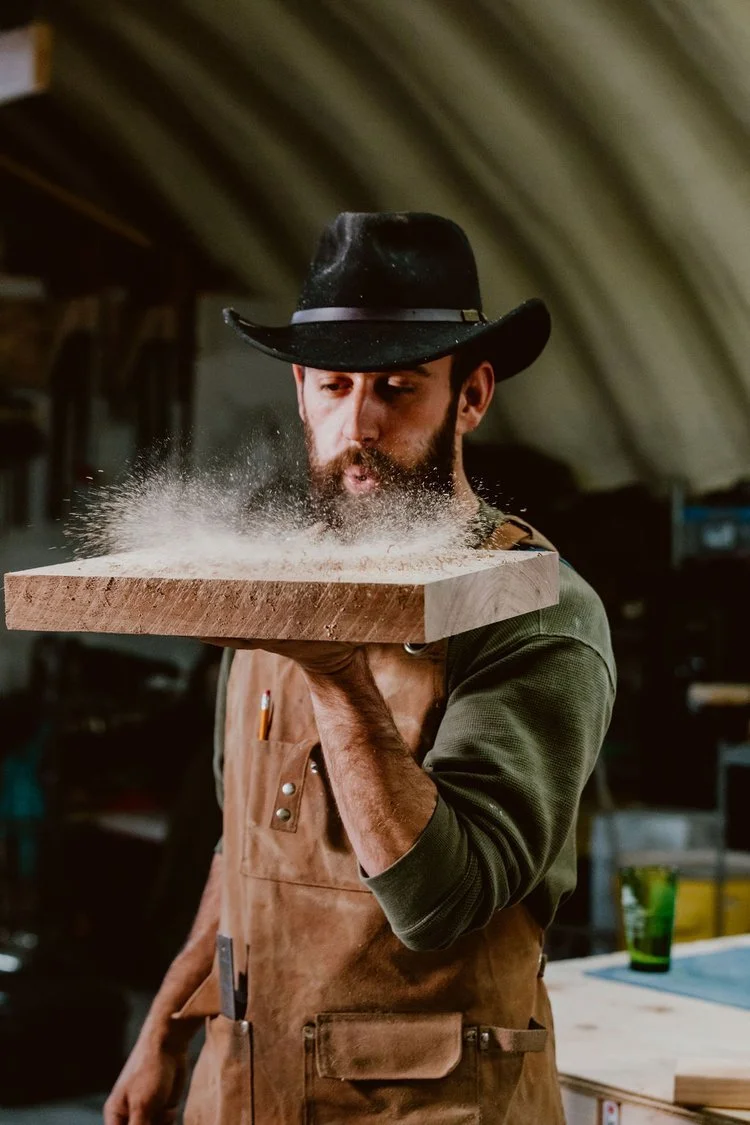 A man wearing a black cowboy hat, green sleeve, and brown apron is standing in a workshop, holding a wooden cutting board close to his face. Dust or powder appears to be falling from the board.