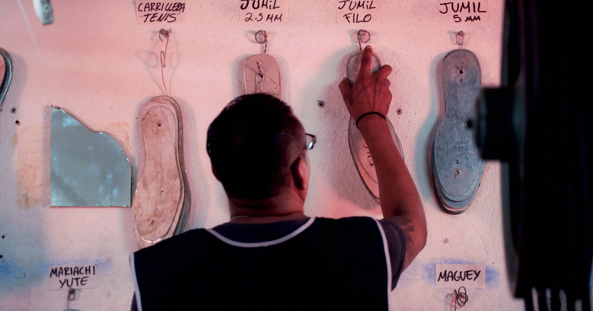A man with glasses and tattoos on his arm points at a silhouette of a shoe on a wall display. The display features various shoe cutouts with labels and measurements, including 'Carrillera Tennis', 'Jumil 2-5 MM', 'Jumil Filo', and others. A small mirror is attached to the wall, and the background wall is painted pink.