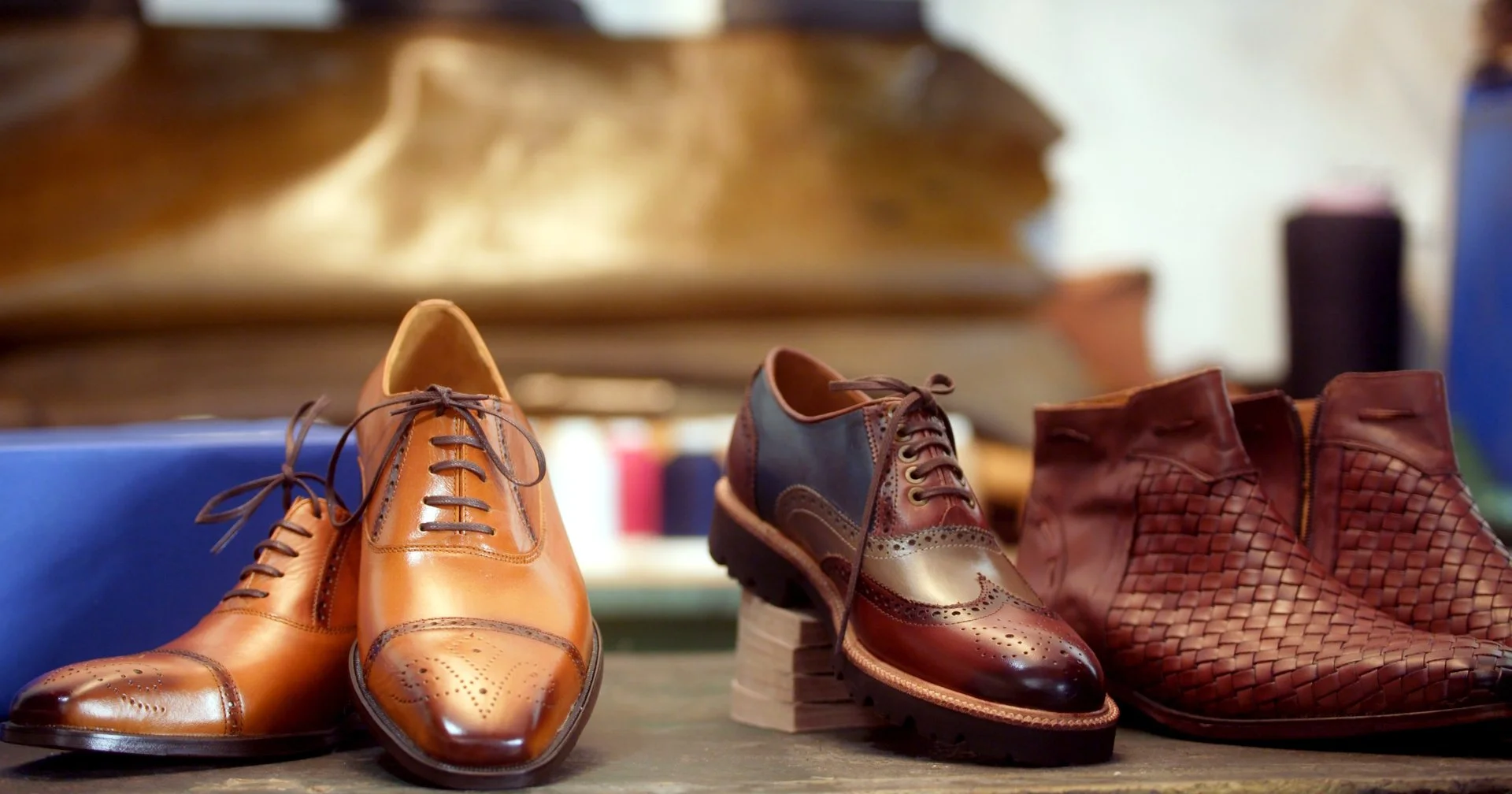 A display of three pairs of men's shoes, including tan leather dress shoes, two-tone brogue shoes, and woven brown leather shoes, on a wooden surface.
