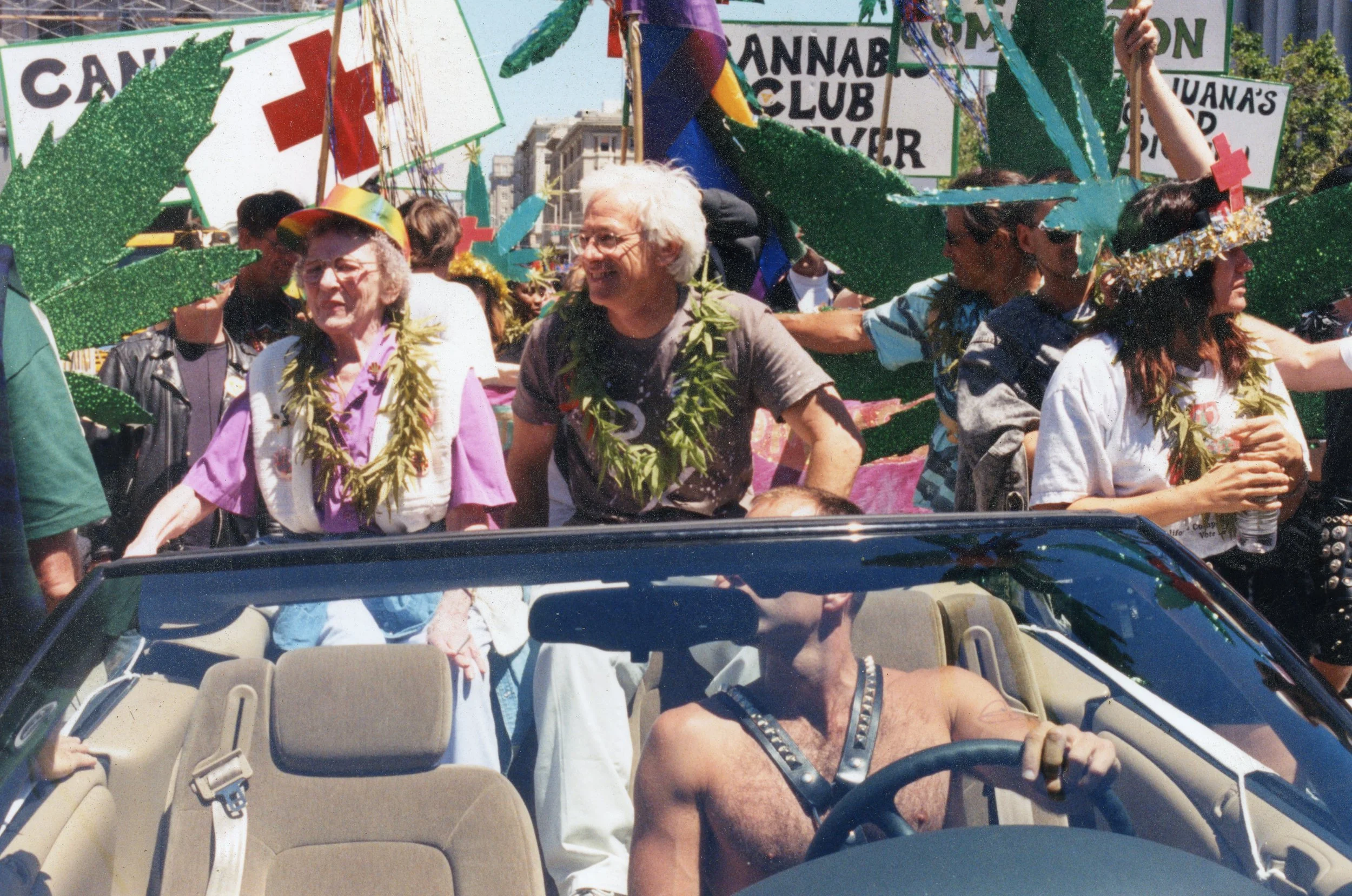 People participating in a parade with signs promoting marijuana and cannabis legalization, riding in a convertible car under bright sunny weather.