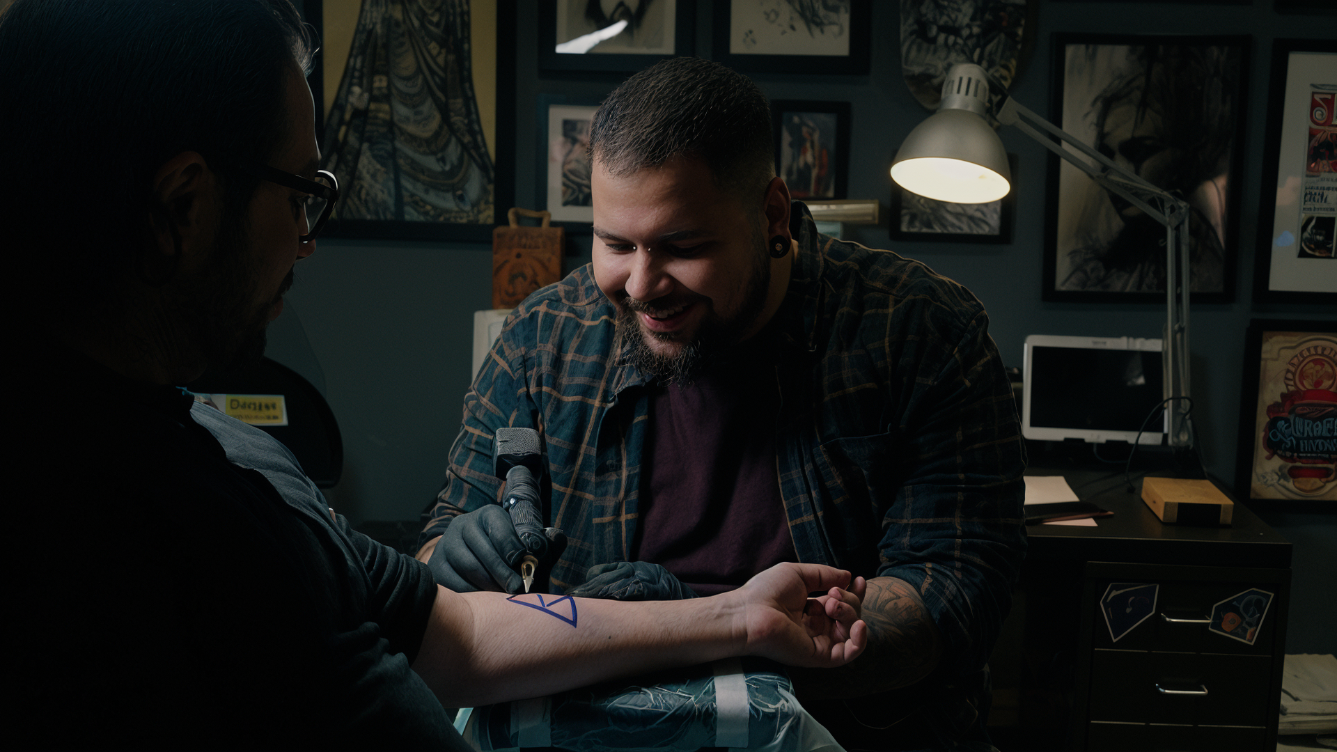 A tattoo artist and a client with a arm tattoo in a tattoo shop, tattooing a blue geometric design.