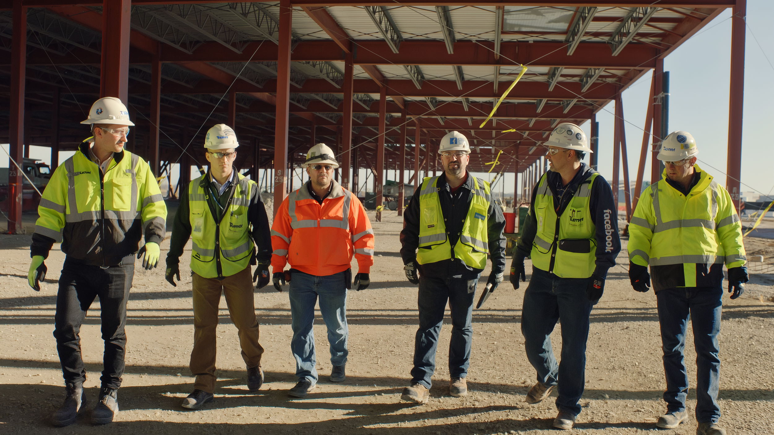 Construction workers wearing safety gear walking on a construction site with a partially built structure overhead.