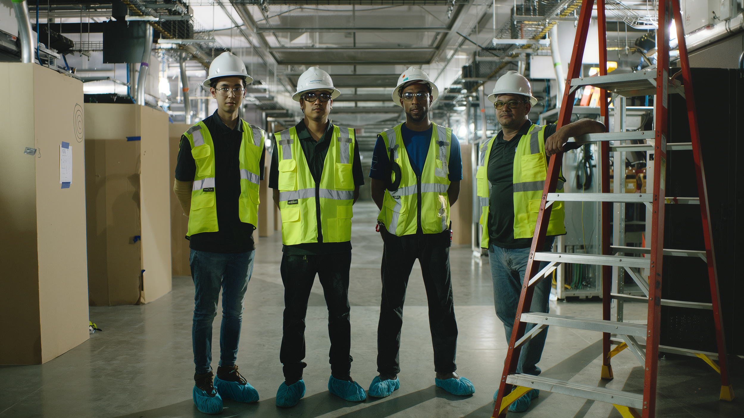 Four workers in safety gear, including helmets and high-visibility vests, standing in an industrial setting with equipment and scaffolding, with shoe covers on their feet.
