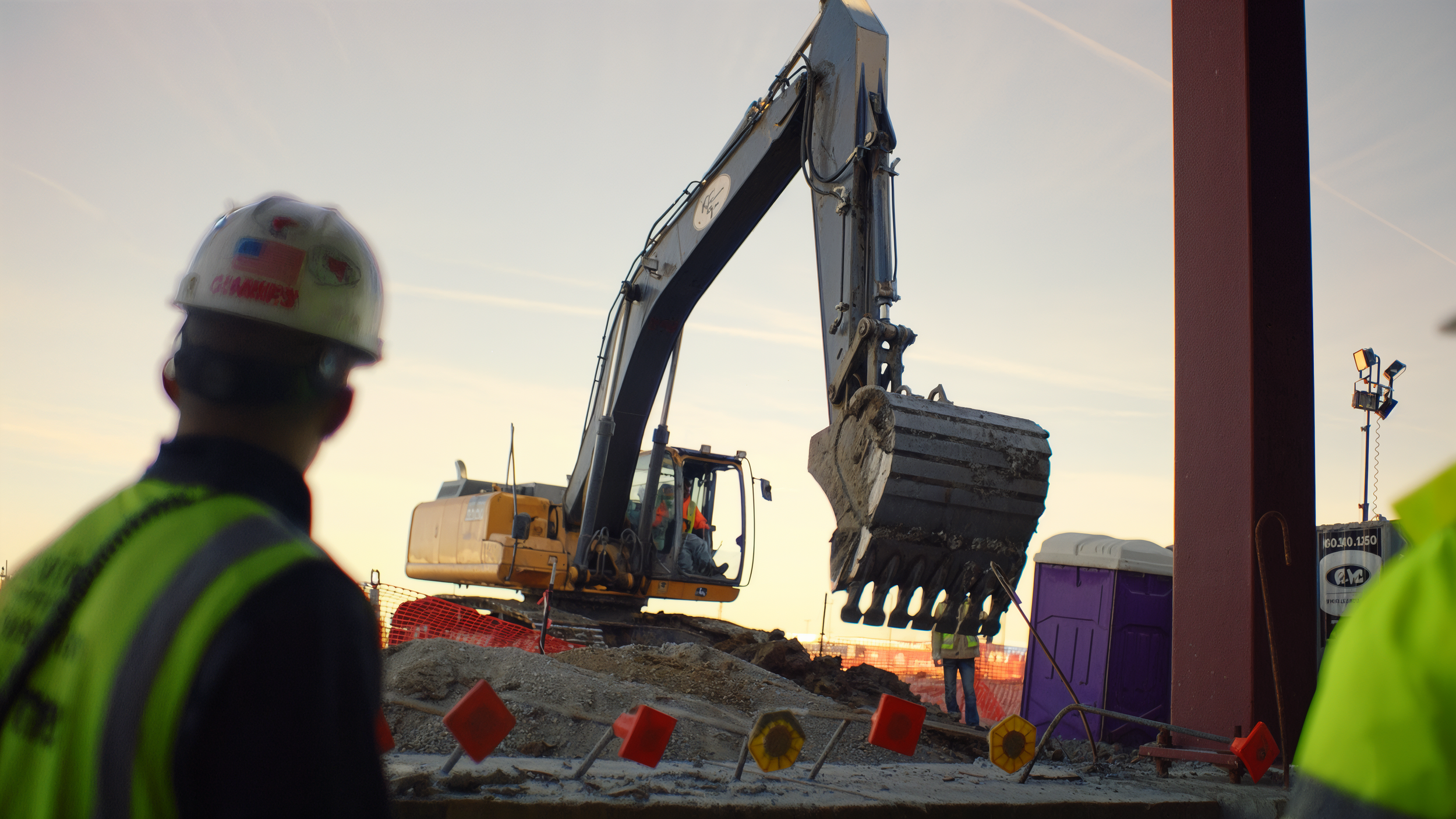 Construction workers wearing high-visibility vests and helmets working on a building site with heavy machinery and orange safety barriers during sunset.