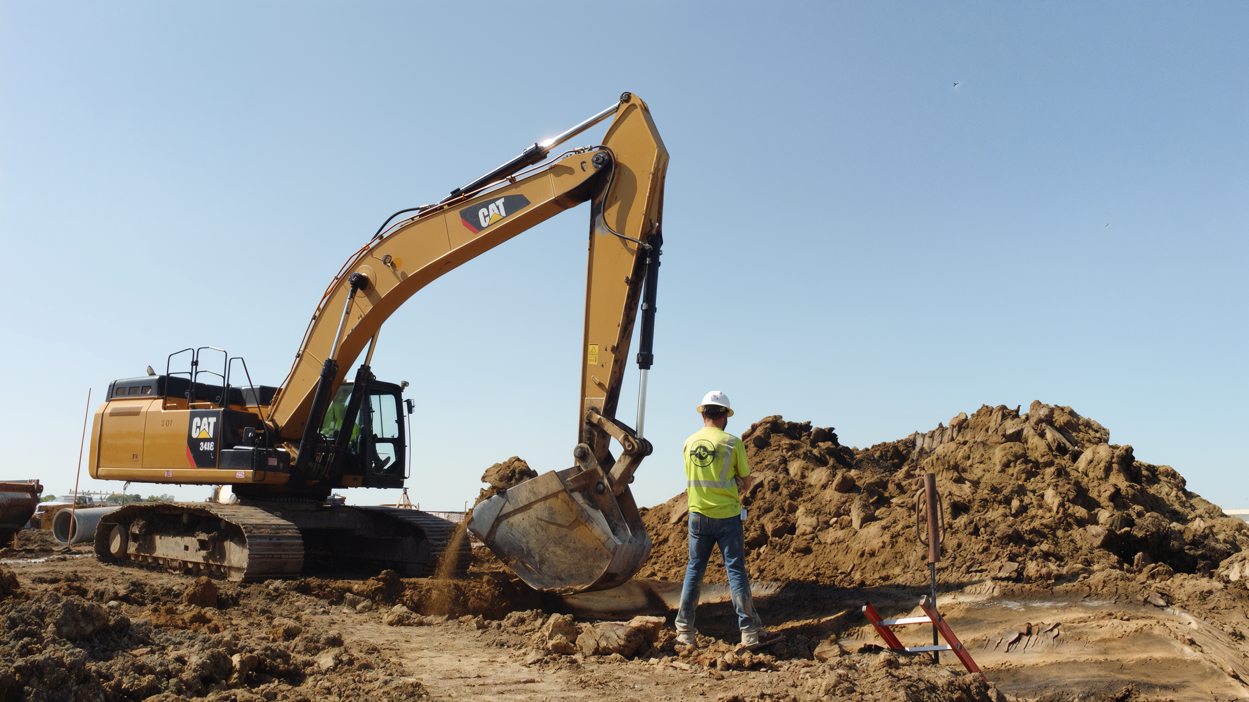 A construction worker in a yellow safety vest and white helmet stands on dirt at a construction site, watching a large yellow Caterpillar excavator digging into a pile of dirt under a clear blue sky.