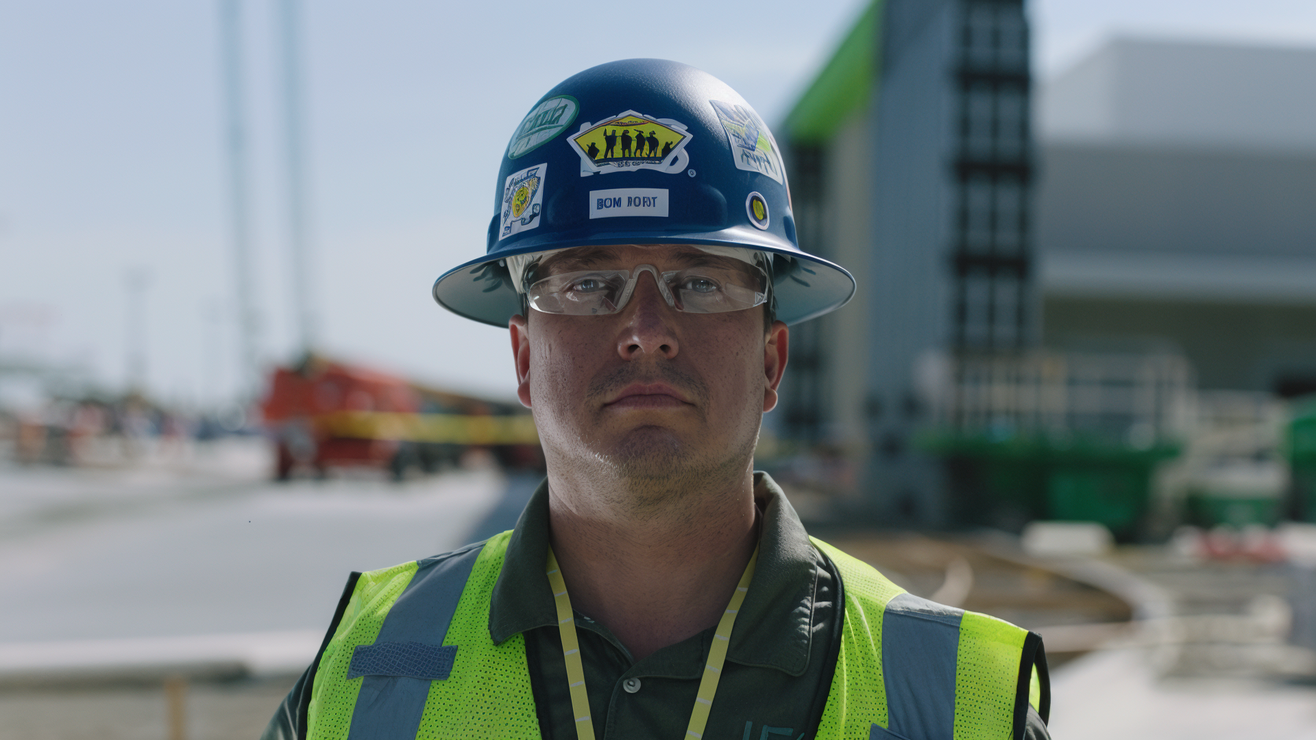 A male construction worker wearing a hard hat with various stickers and clear safety glasses at a construction site.