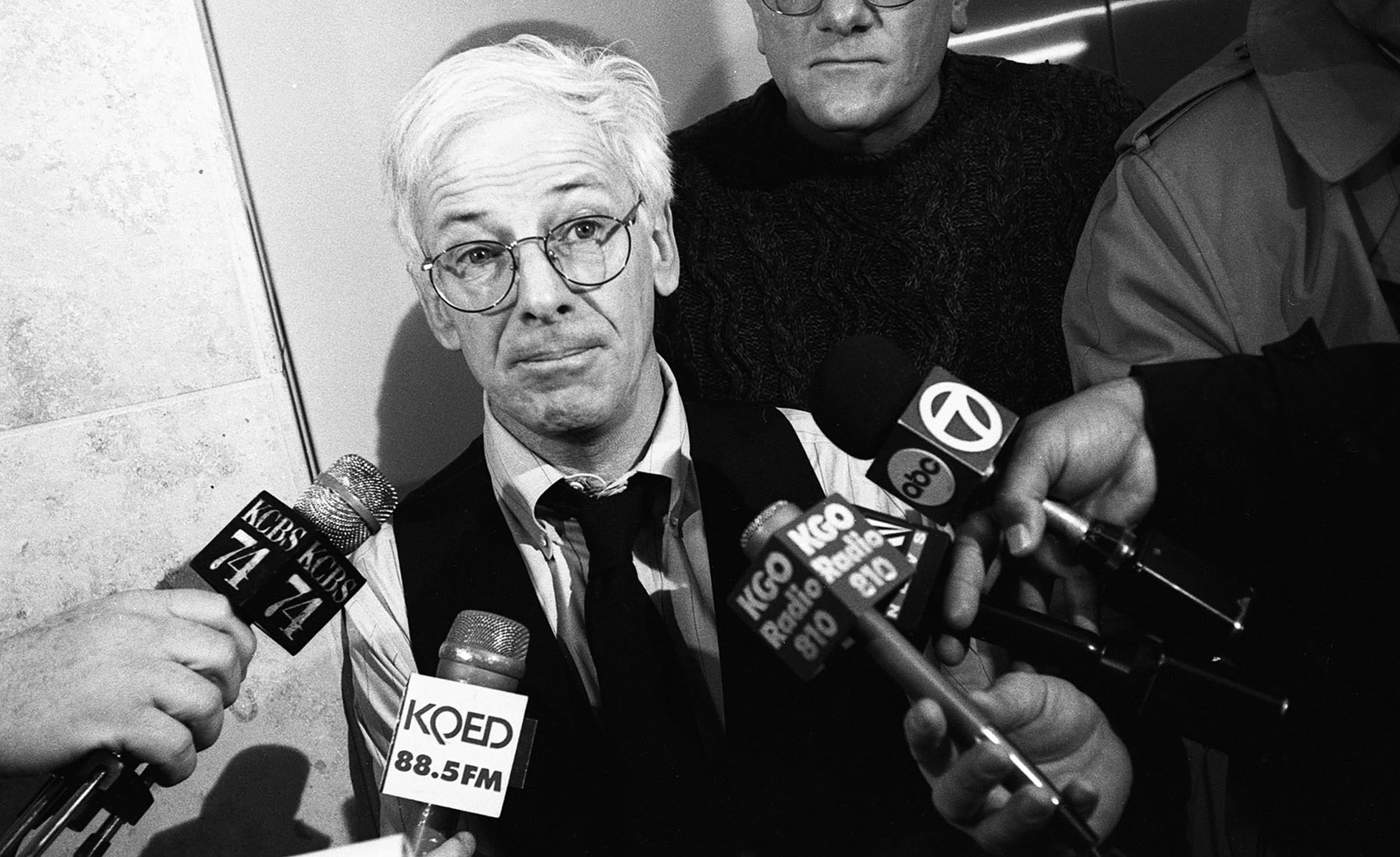 Black and white photo of a person with glasses and light-colored hair speaking to reporters, multiple microphones labeled with radio station logos are directed at him, and several hands hold recording devices close to his face.