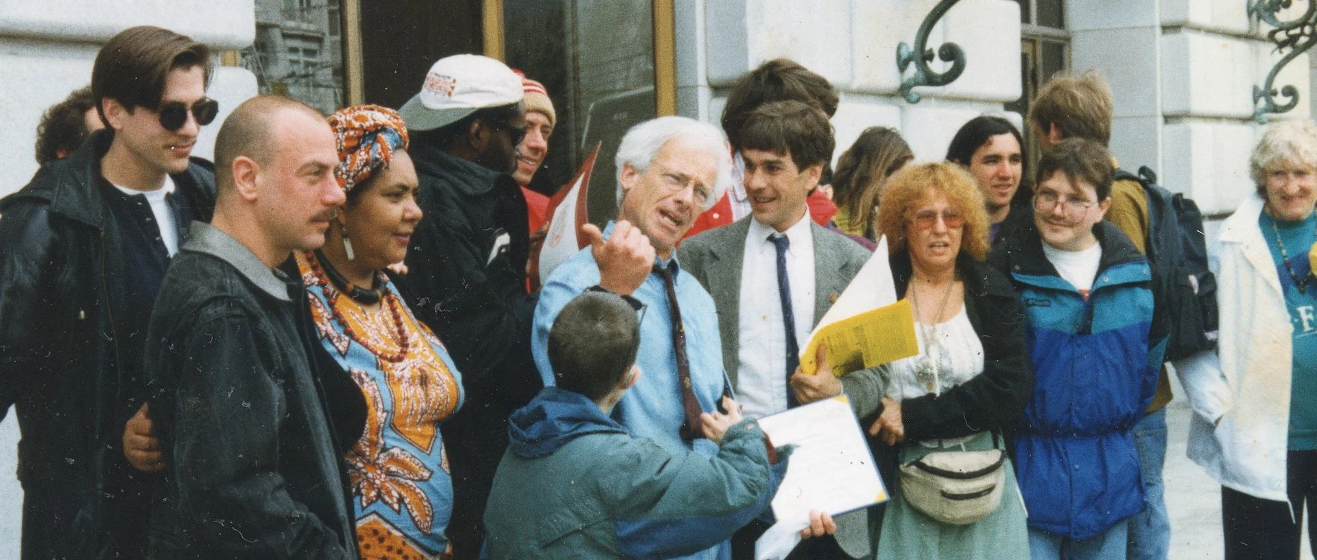 Group of diverse people gathered outdoors, some holding papers and others listening, engaged in a conversation.