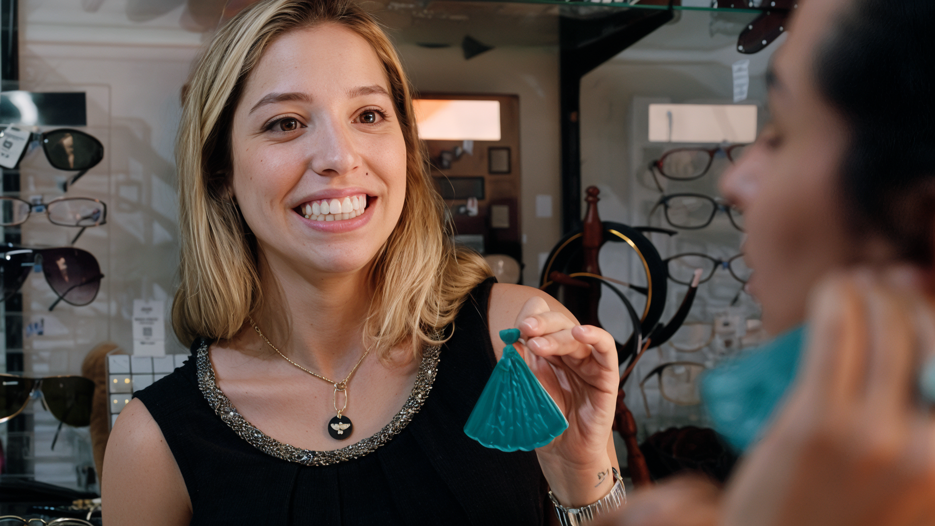 A woman smiling and showing her teeth as she hands a small blue bag to a person in a store that sells glasses, with shelves of glasses in the background.