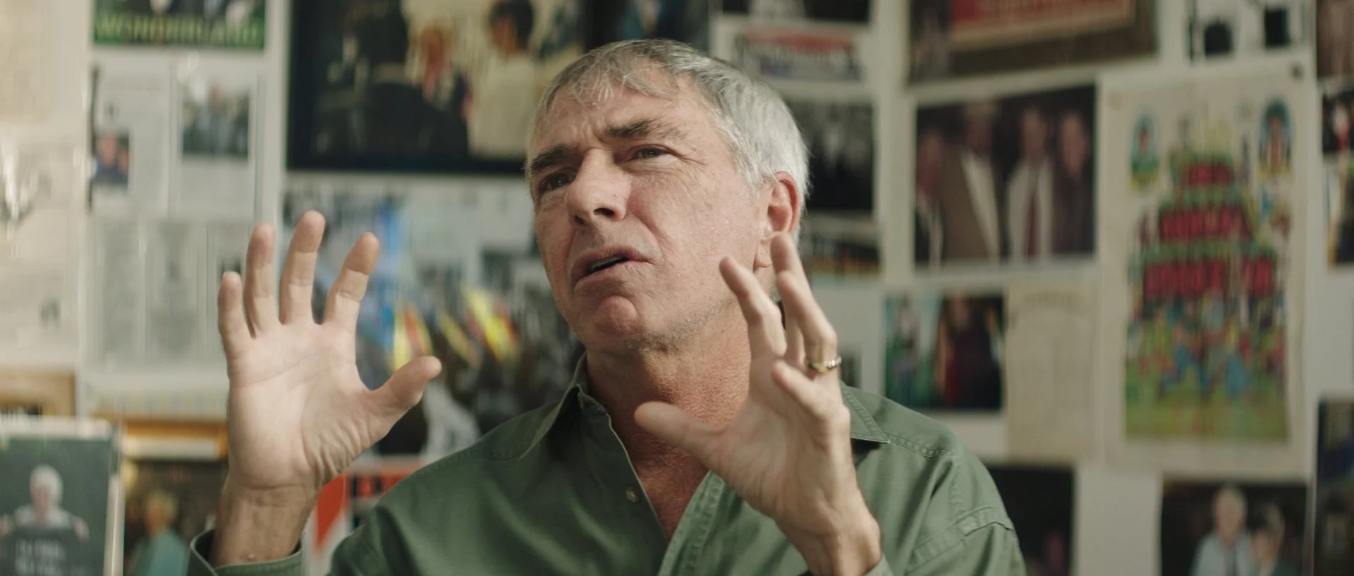 A middle-aged man with gray hair, wearing a green shirt, appears to be talking passionately with his hands raised, in a room decorated with photographs and posters on the walls.