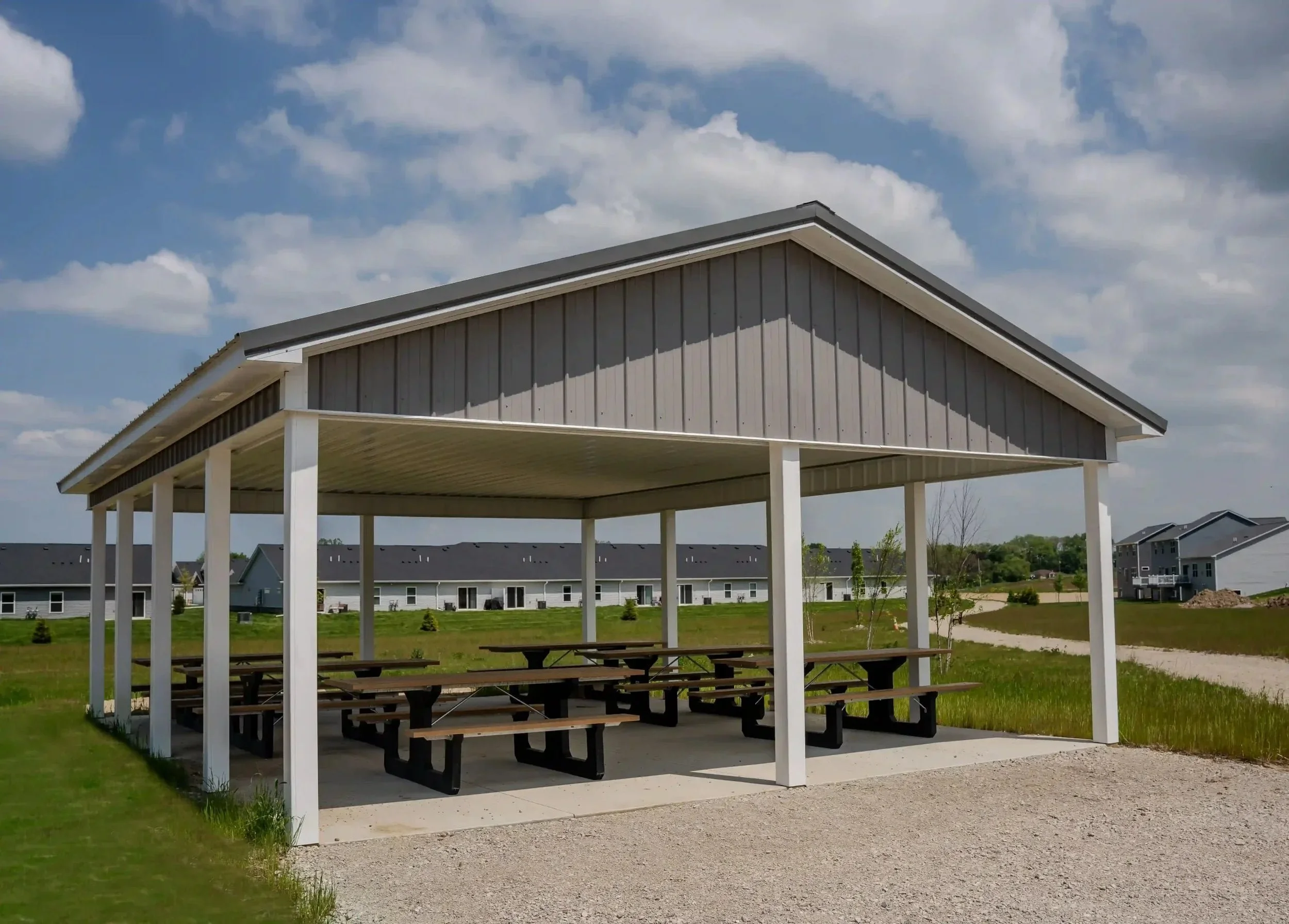 A pavilion with a roof supported by white columns, featuring picnic tables underneath, situated on a grassy area with residential buildings and a blue sky with clouds in the background.
