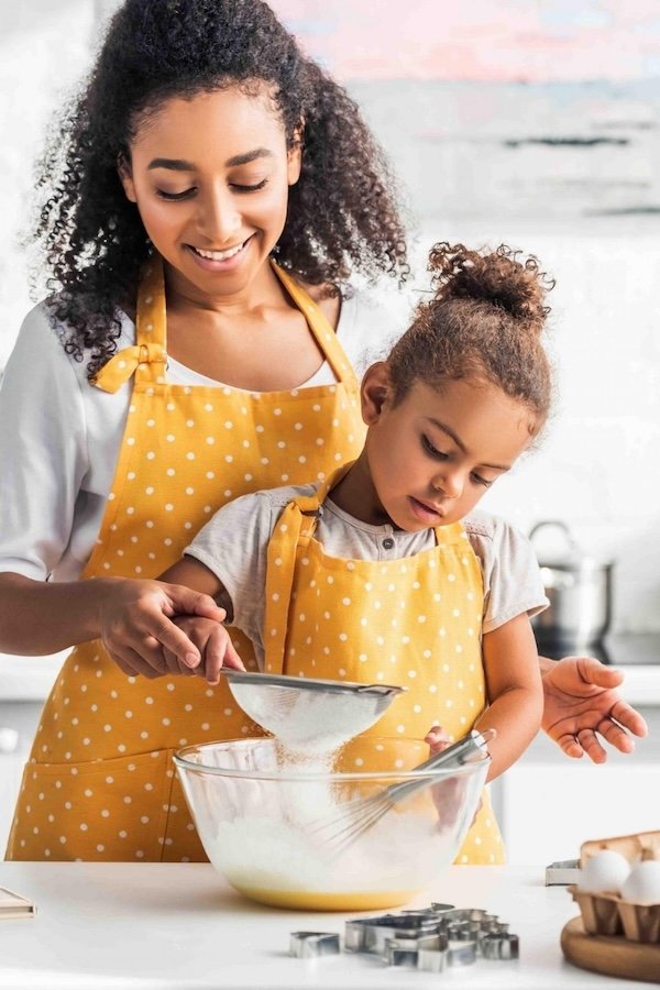 A woman and young girl baking together in the kitchen, both wearing yellow polka dot aprons, with the woman helping the girl sift ingredients into a bowl.