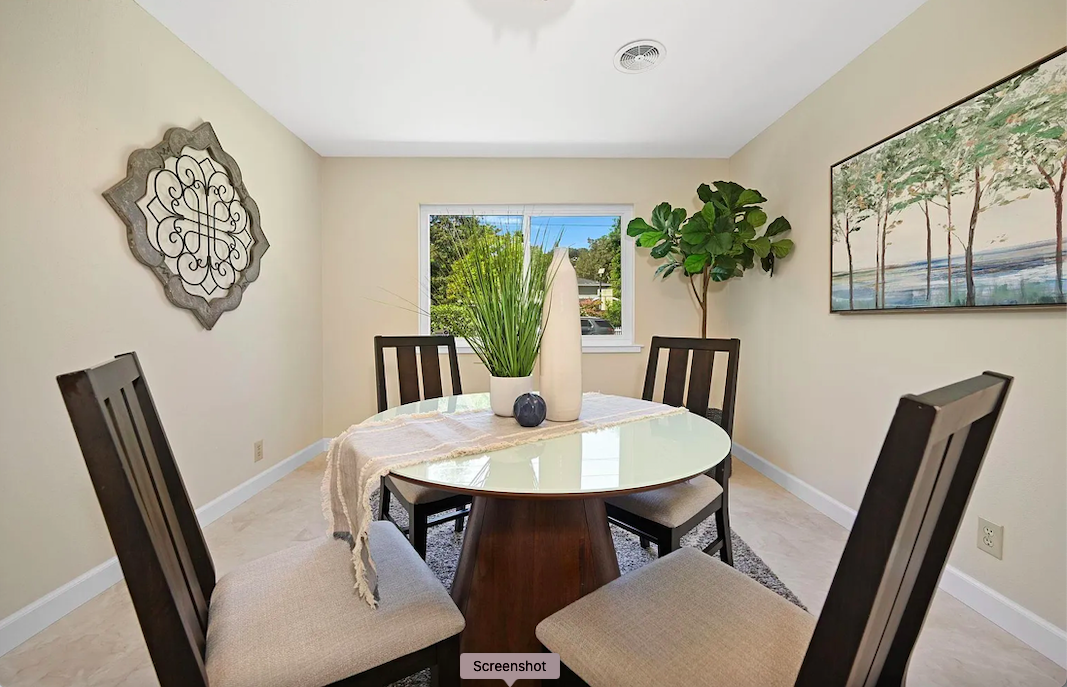 Dining room with a round glass table, four dark wood chairs with beige cushions, a window, a large potted plant, a decorative wall element, and a landscape painting on the wall.