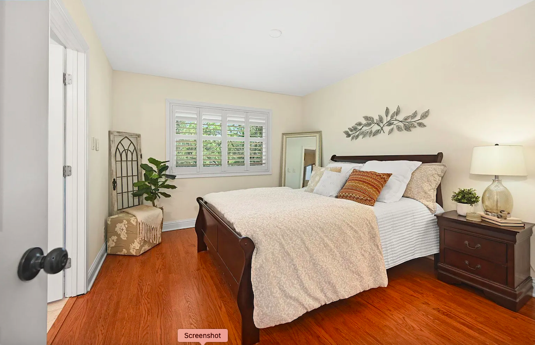 A cozy bedroom with a wooden bed, white bedding, decorative pillows, a nightstand with a lamp and plant, a full-length mirror, and a window with shutters.