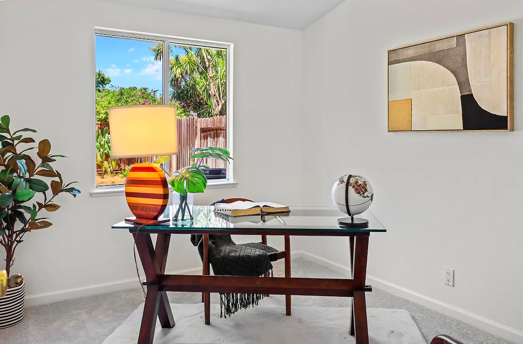 Home office with a glass desk, a chair, a table lamp, plants, a globe, and a window showing greenery outside.