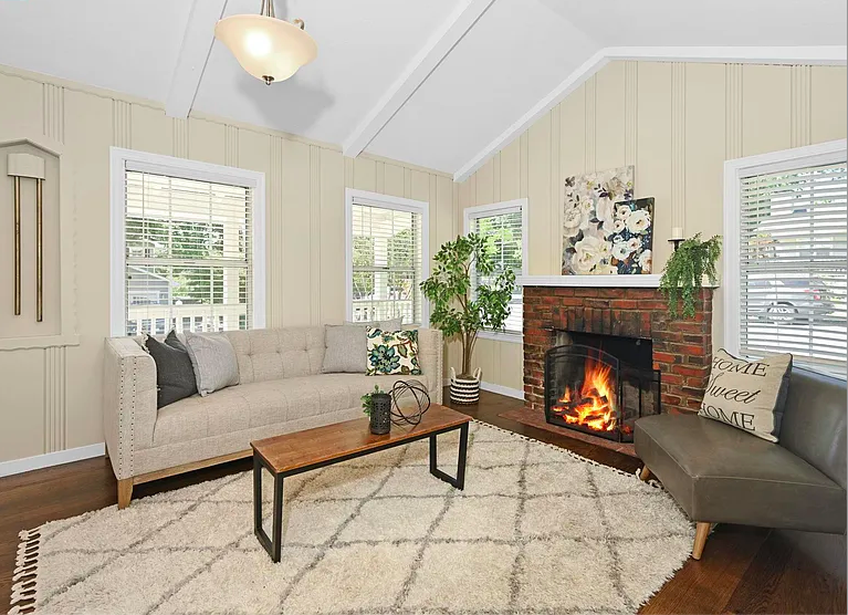 Living room with beige walls, white trim, four windows with blinds, a brick fireplace with a fire, a gray sofa with pillows, a gray side chair with a pillow, a wooden coffee table, a large white rug, and decorative plants.