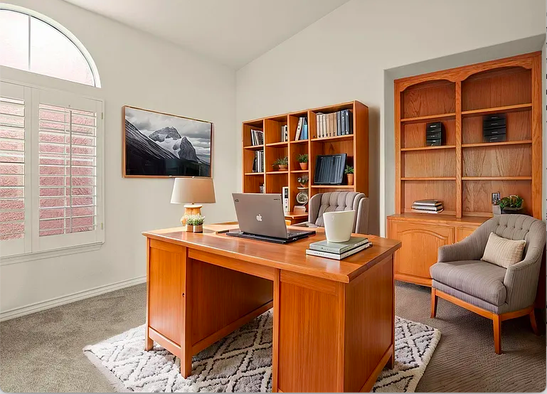 Home office with a wooden desk, a laptop, a lamp, and a chair, with bookshelves and a window with shutters.