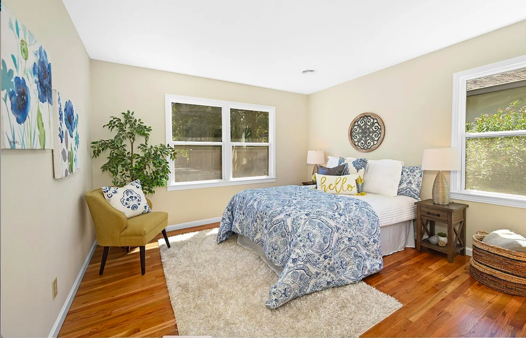 A bedroom with beige walls, hardwood floors, a bed with blue and white bedding, two bedside tables with lamps, a yellow accent chair with a patterned pillow, a large window, wall art, a throw pillow with "hello" written on it, a white shag rug, and a