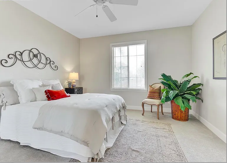 Bright bedroom with a white bed, decorative metal wall art, a bedside lamp, a window with blinds, a wooden chair, a large green potted plant, and a framed picture on the wall.
