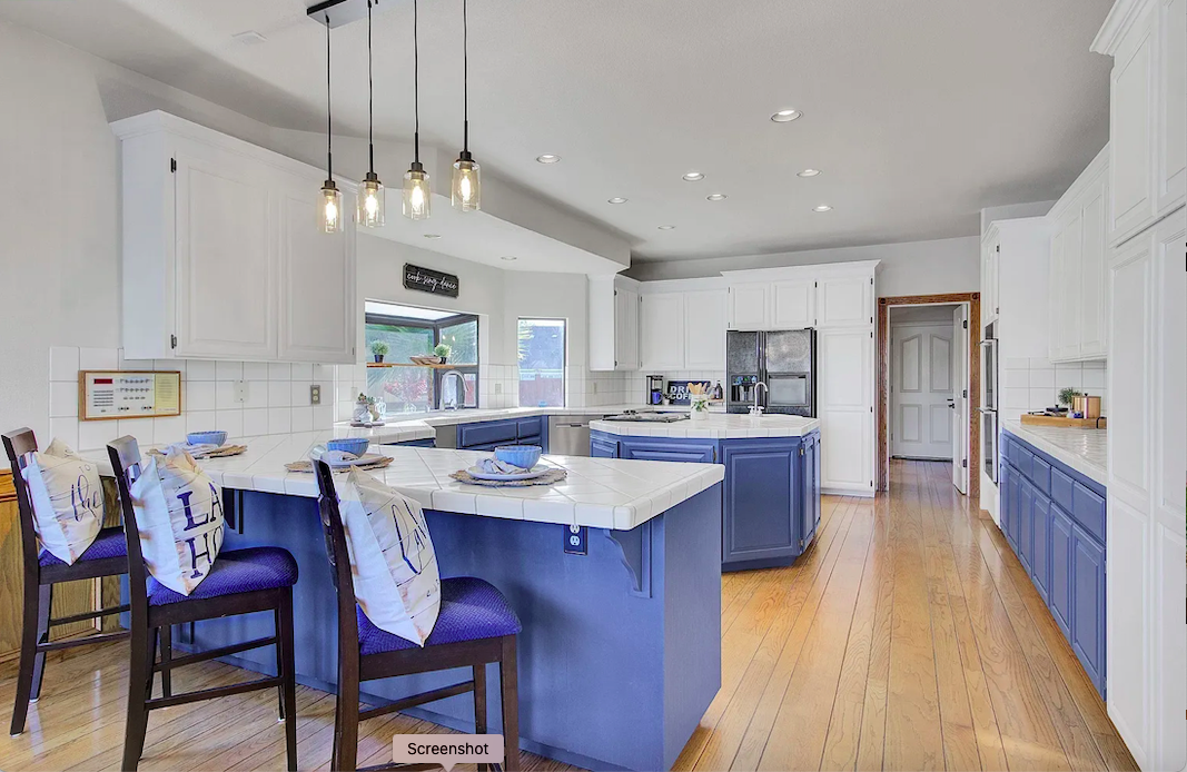 Kitchen with white cabinets, blue island, wooden flooring, pendant lights, and bar stools with pillows.