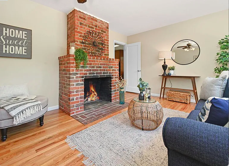 Living room with a red brick fireplace, a built-in fire, cream-colored walls, a gray loveseat with throw pillows, a wicker coffee table with decorative vases, a gray armchair with a blanket, a wooden side table with a lamp, a round mirror, a wall sig