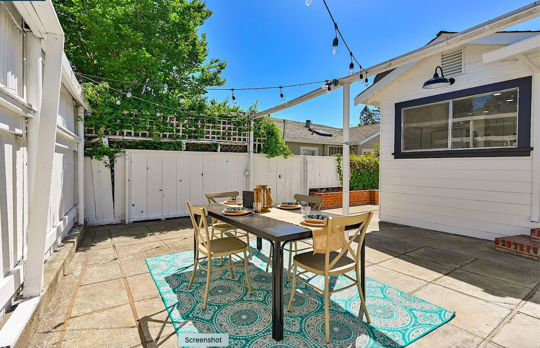 Outdoor patio with a dining table set for four under a string of outdoor lights, on a patterned rug, surrounded by a white fence and a house with a large window and black trim.