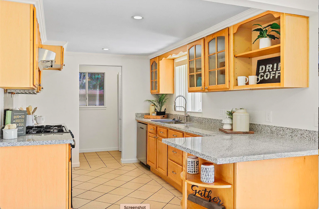 Kitchen with wooden cabinets, granite countertops, and a window above the sink. Decor includes potted plants, mugs, and jars, with a tiled floor.