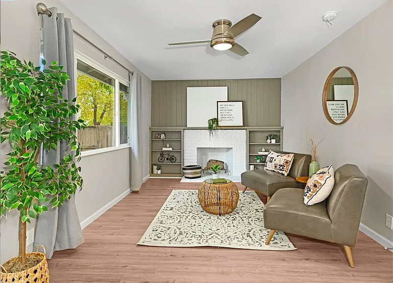 Living room with a large window, gray curtains, a potted plant, a fireplace with built-in shelves, a decorative mirror on the wall, a beige rug, a round wicker coffee table, and two gray upholstered chairs with patterned pillows.