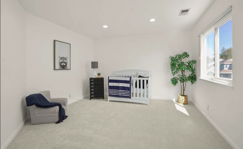 A minimalist nursery with white walls, carpet, a white crib with navy blanket, a small black dresser with a lamp, a picture of a dog on the wall, a gray armchair with a blue throw, a large green potted plant, and a window showing a neighborhood outsi