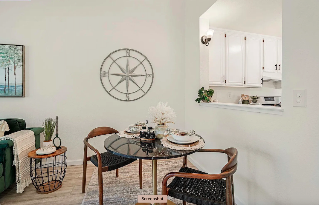 A dining area with a black round table, surrounded by three wooden chairs with black woven seats. On the table are decorative bowls, a white dried flower centerpiece, and placemats. To the left, there's a green armchair with a white throw blanket and