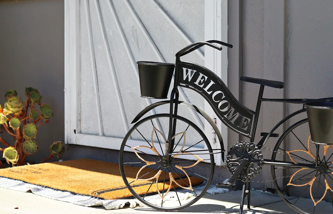 Decorative bicycle planter with a welcome sign, black flower pots, placed on a porch with a doormat and a potted succulent plant nearby.