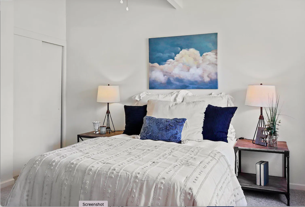A neatly made bed with white bedding, blue pillows, and a blue cushion, flanked by two nightstands with lamps. Above the bed hangs a painting of clouds in a blue sky. The room has bright lighting and minimal decor.