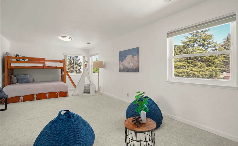 Bright children's bedroom with white walls and carpet, featuring a wooden bunk bed, a teepee tent, a large window showing trees outside, and cozy seating with blue poufs and a small round table.