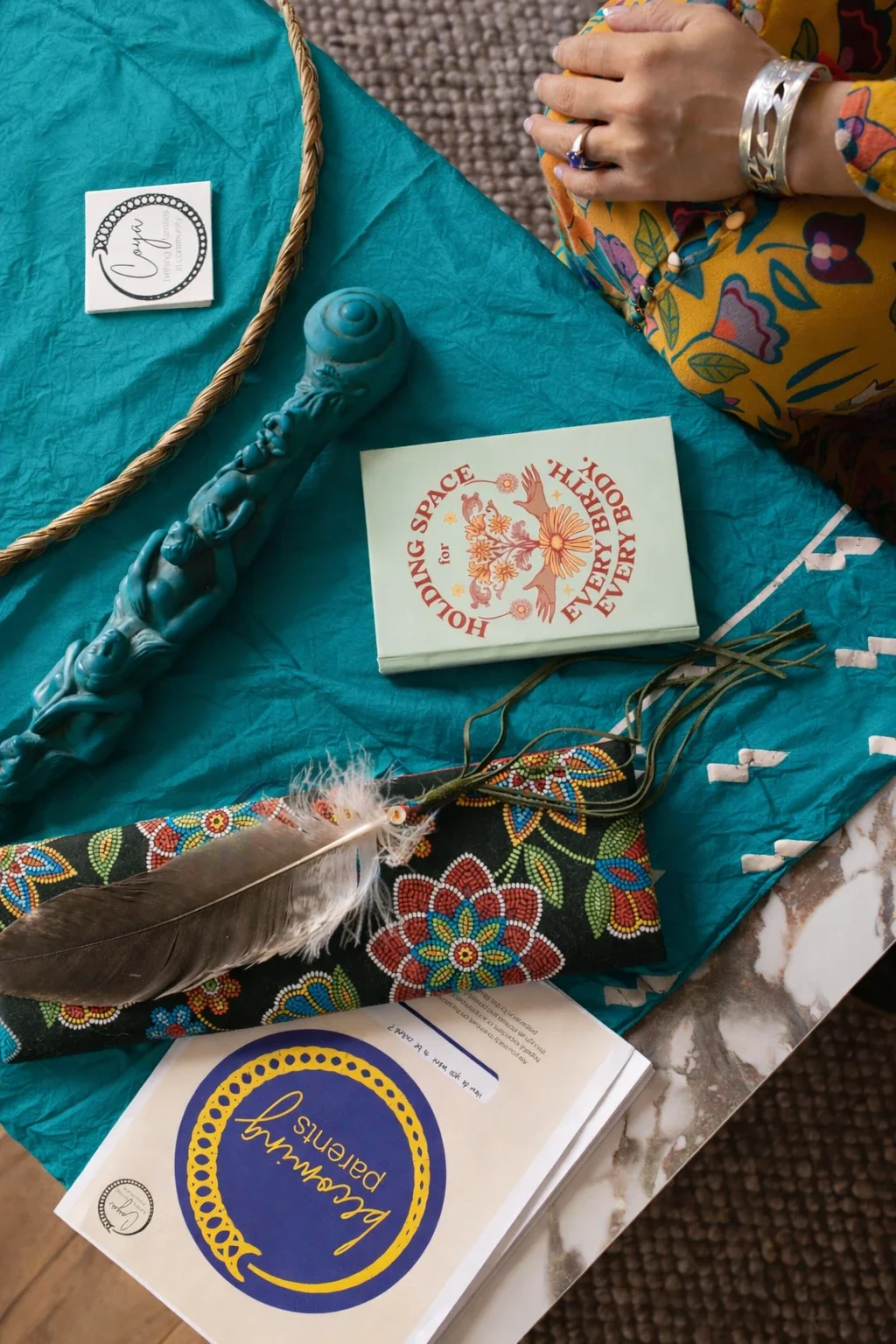 Arrangement of various items on a table including a light blue candle with an intricate design, a booklet titled 'HOLDING SPACE for EVERYBODY'S DAY, EVERYBODY'S BODY,' a Feathered accessory, and a card with the words 'Breakfast & Pains' inside a blue and yellow circle. A person's hand resting on a colorful floral sleeve is visible in the upper right corner.