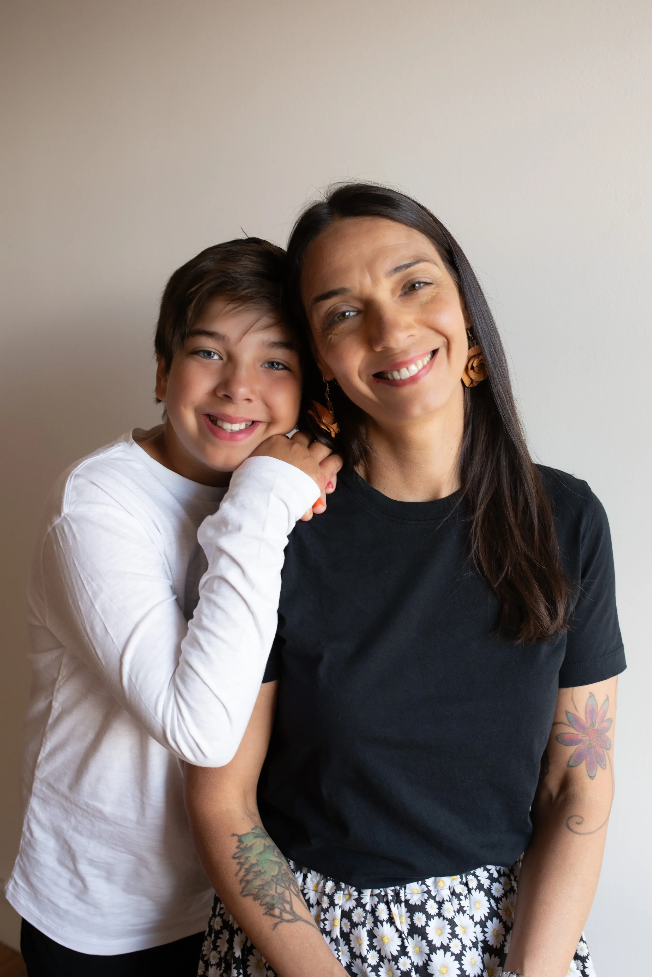 A woman smiling with a young boy leaning on her shoulder, both looking at the camera, standing against a plain light-colored wall.