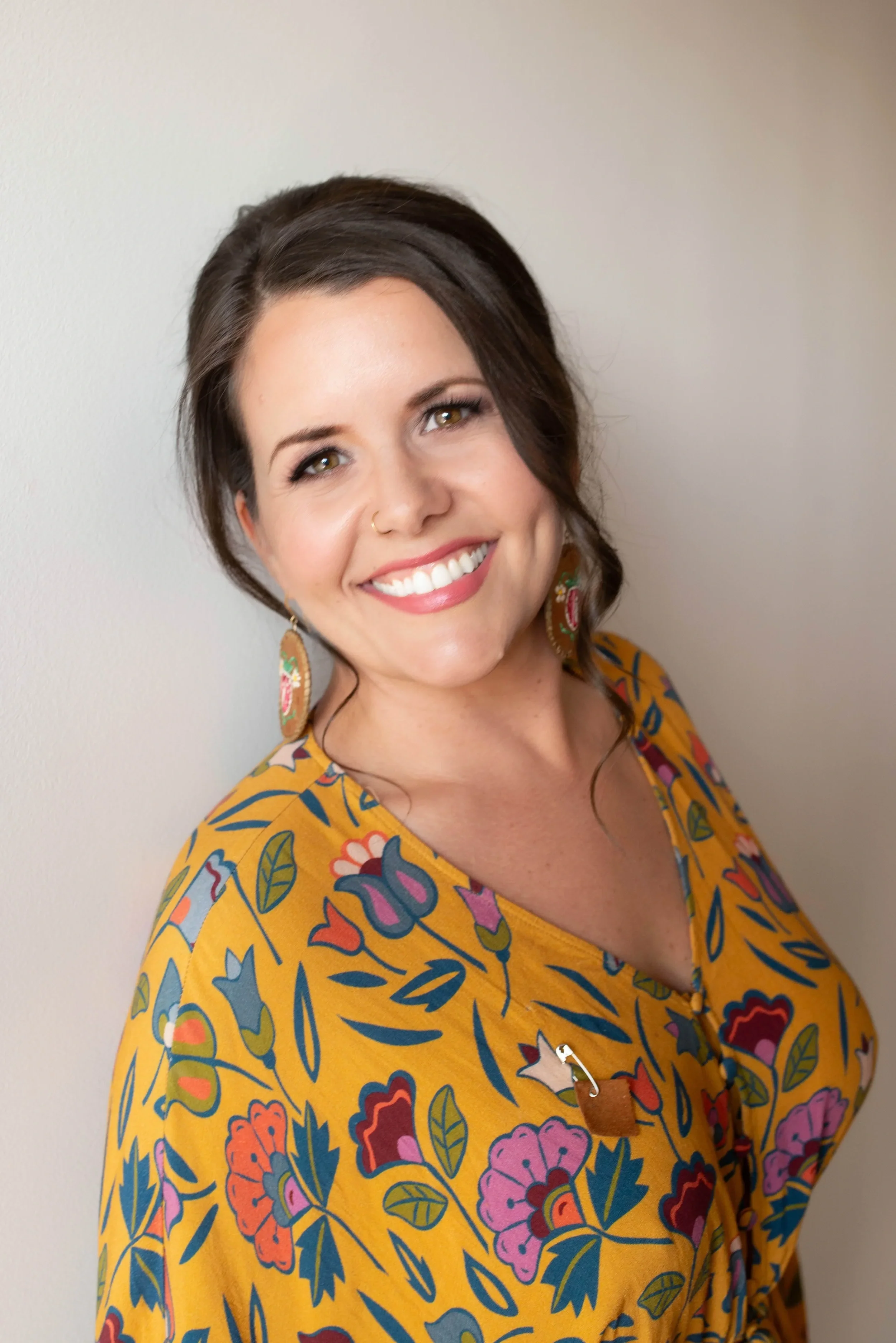 A woman with dark brown hair, smiling, wearing colorful floral earrings and a yellow blouse with a vibrant floral pattern, standing against a plain wall.