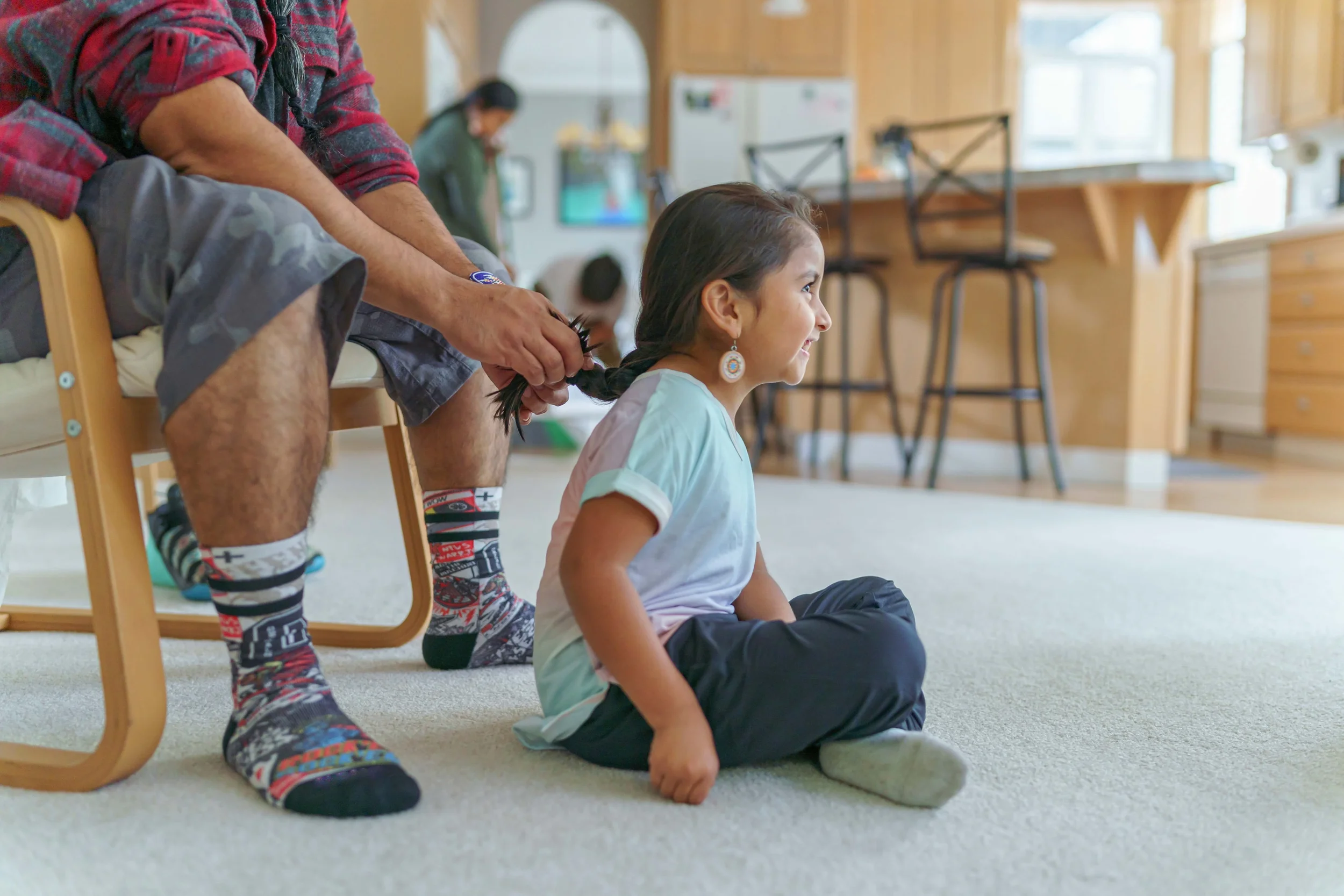 A man is sitting on a chair, braiding a young child's hair. The child is sitting on the floor with her legs crossed, smiling and looking to the side.